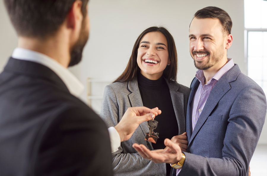 Close up shot of happy hugging young couple receiving new home keys from real estate agent. Professional male realtor handing keys and congratulating smiling man and woman with property purchase.