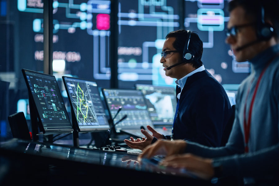 Portrait of Professional IT Technical Support Specialist Working on Computer in Monitoring Control Room with Digital Screens. Employee Wears Headphones with Mic and Talking on a Call.